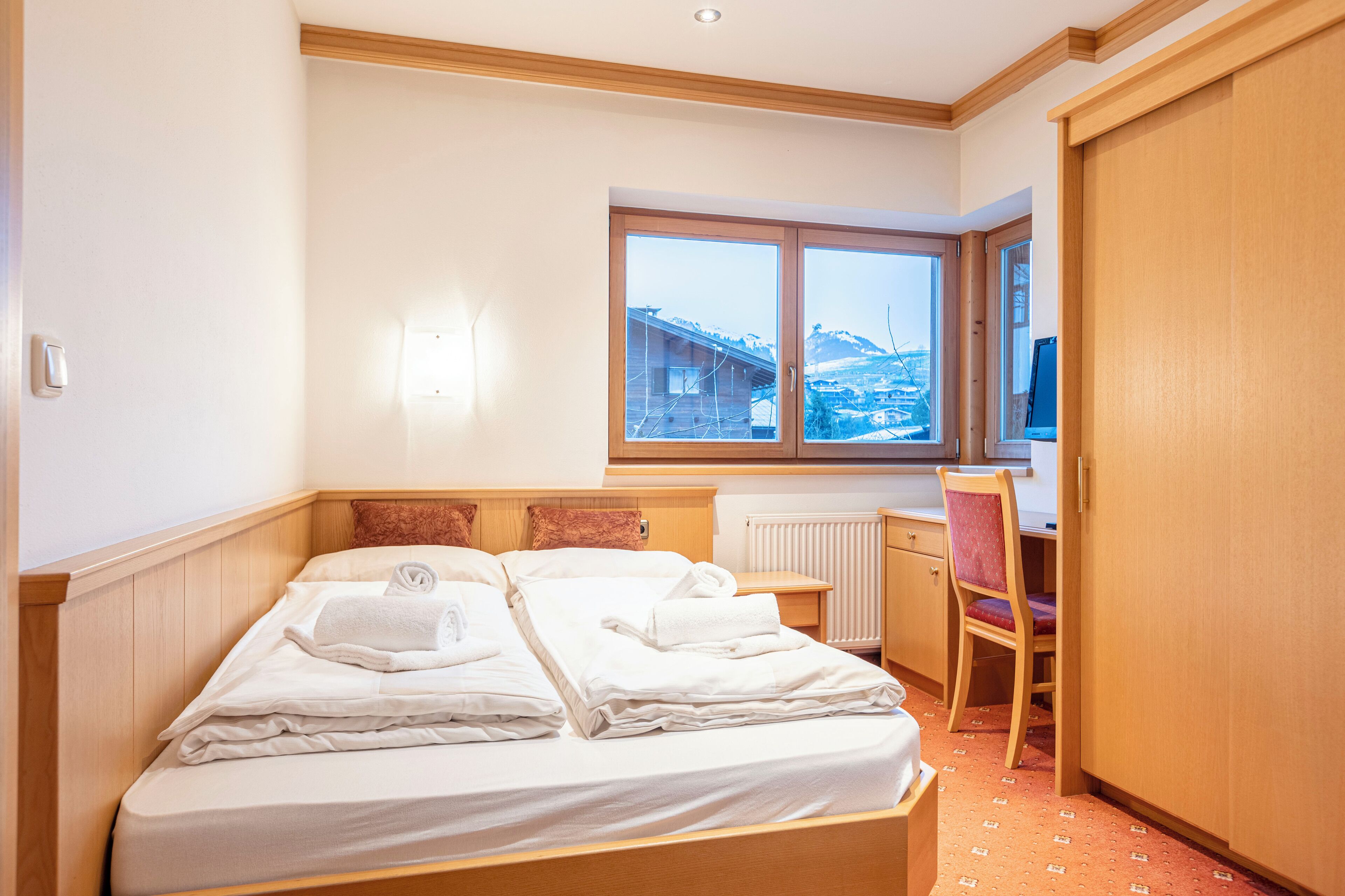 A hotel room with a bed, a desk, a chair, and a window with a view of buildings and mountains.