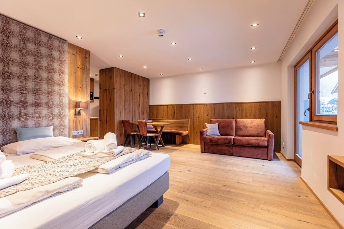A modern hotel room with a large bed, a wooden headboard, a sofa, a dining table, and a large window.
