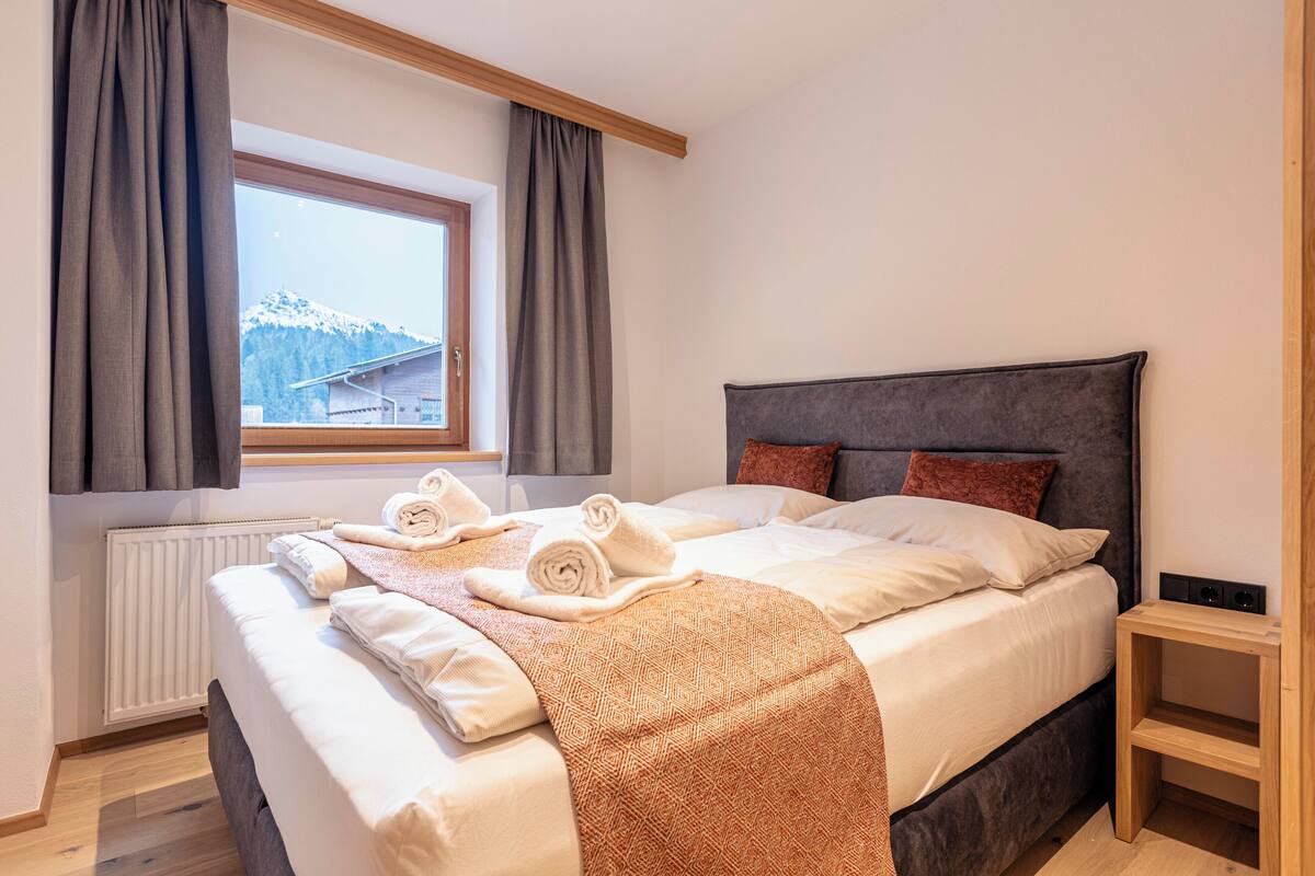 A neatly made bed with towels, a window with curtains, and a view of mountains.