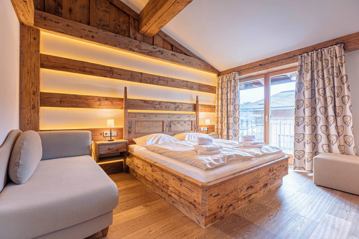 A cozy bedroom with a wooden headboard, a bed with white linens, a nightstand, a bedside table, a sofa, and a window with curtains.