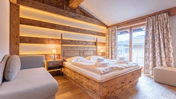 A cozy bedroom with a wooden headboard, a bed with white linens, a nightstand, a bedside table, a sofa, and a window with curtains.