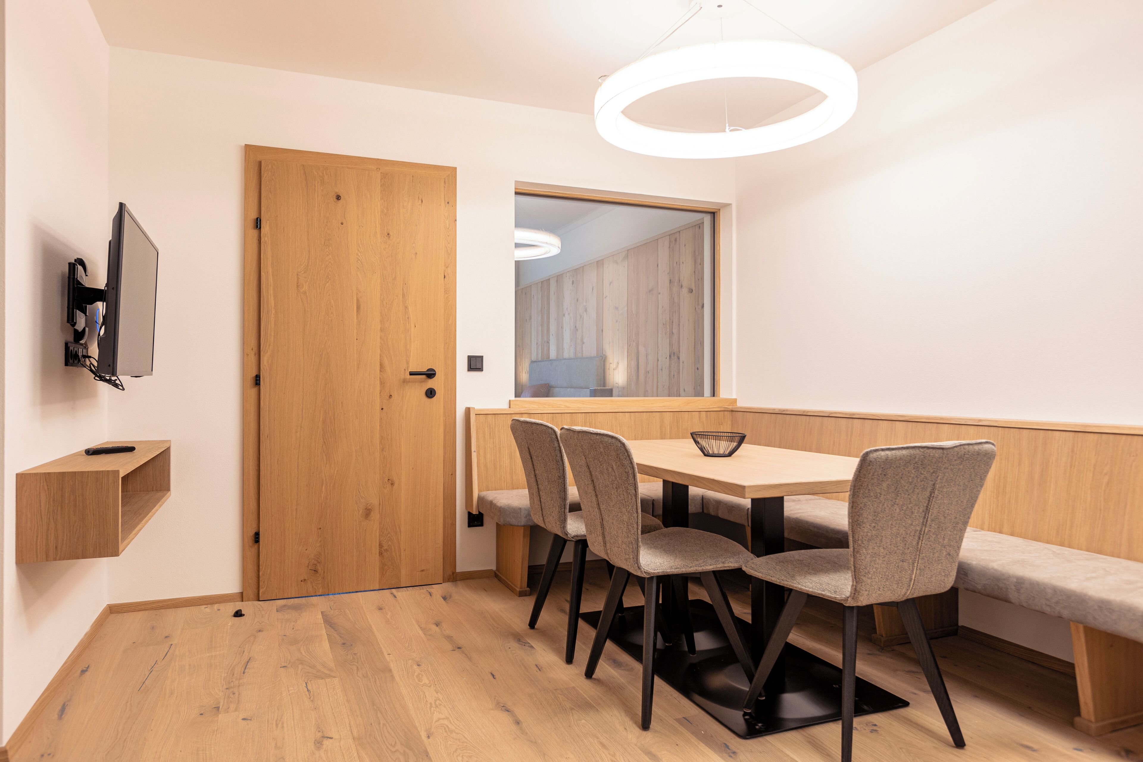 A modern dining area with a wooden table, four chairs, a bench, and a television mounted on the wall.