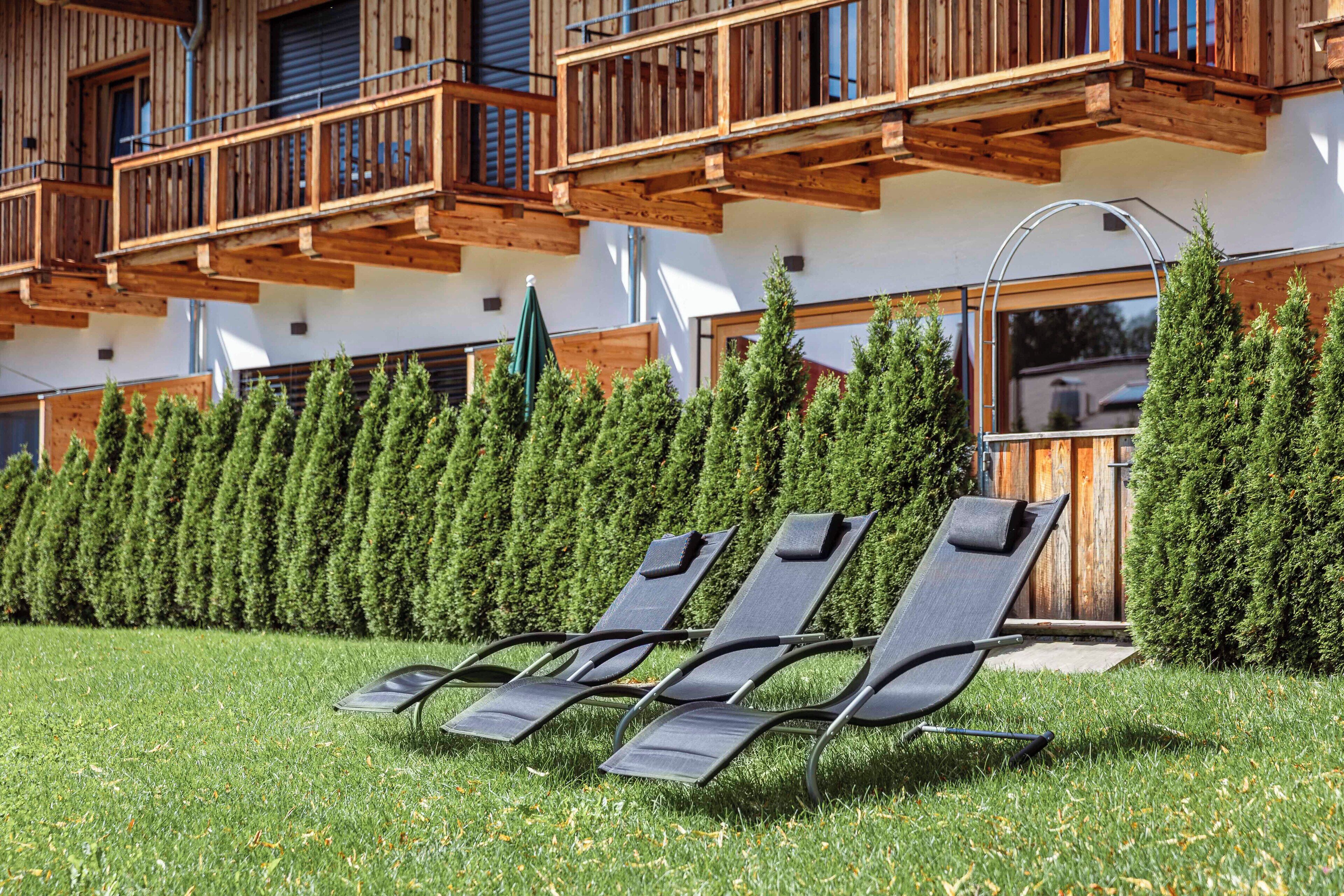 Una hilera de tumbonas en un césped frente a un edificio con balcones de madera y una fila de árboles de hoja perenne.