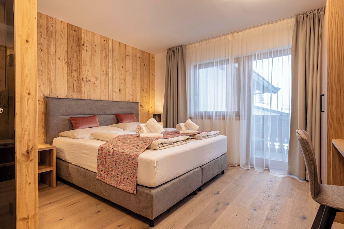 A bedroom with a wooden wall, a bed with pillows and a blanket, a chair, and a window with curtains.