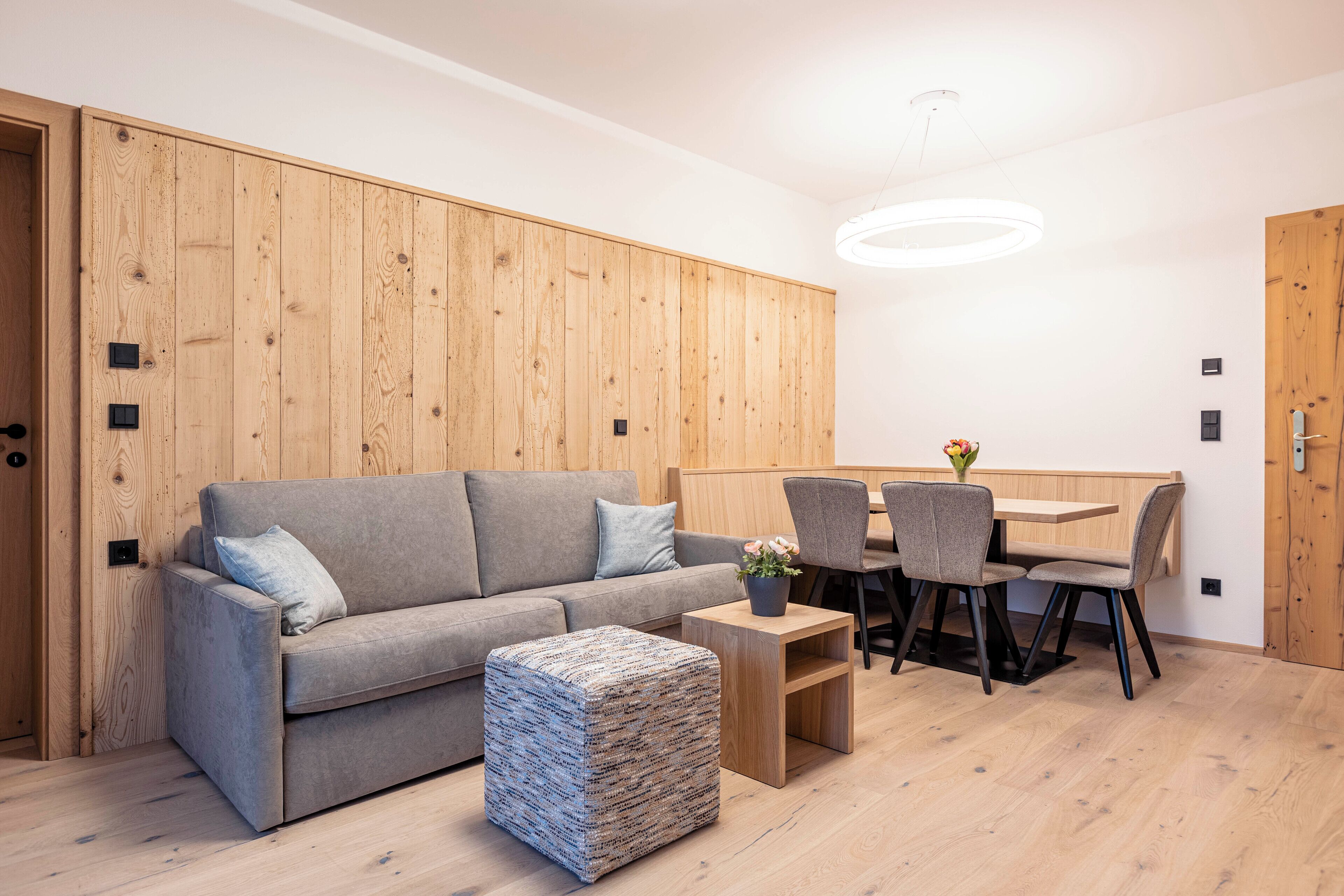 A modern living room with a grey sofa, a small ottoman, a wooden dining table with chairs, and a wooden accent wall.