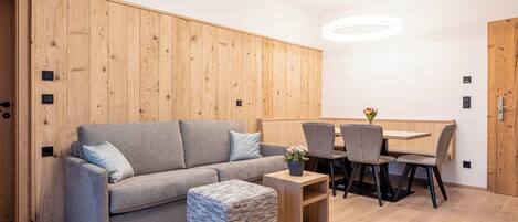 A modern living room with a grey sofa, a small ottoman, a wooden dining table with chairs, and a wooden accent wall.