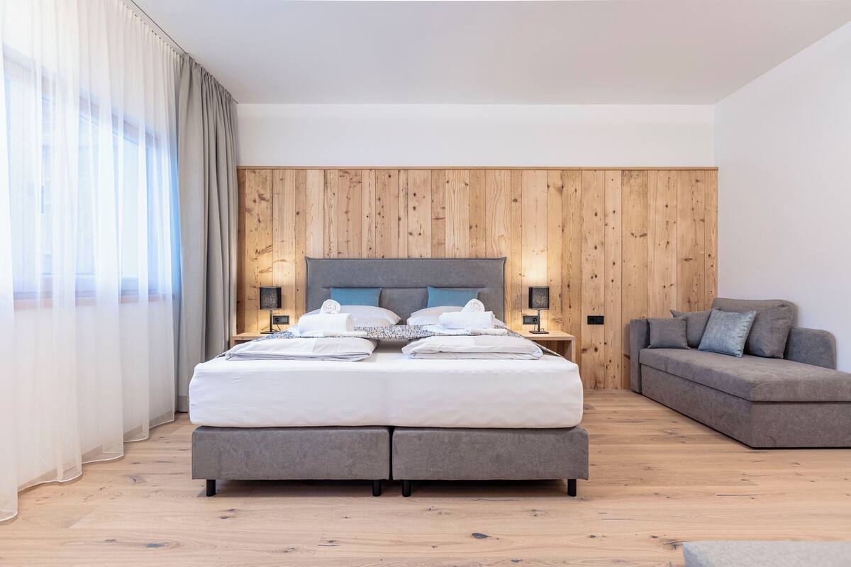 A modern bedroom with a large bed, a grey sofa, wooden paneling on the wall, and a wooden floor.