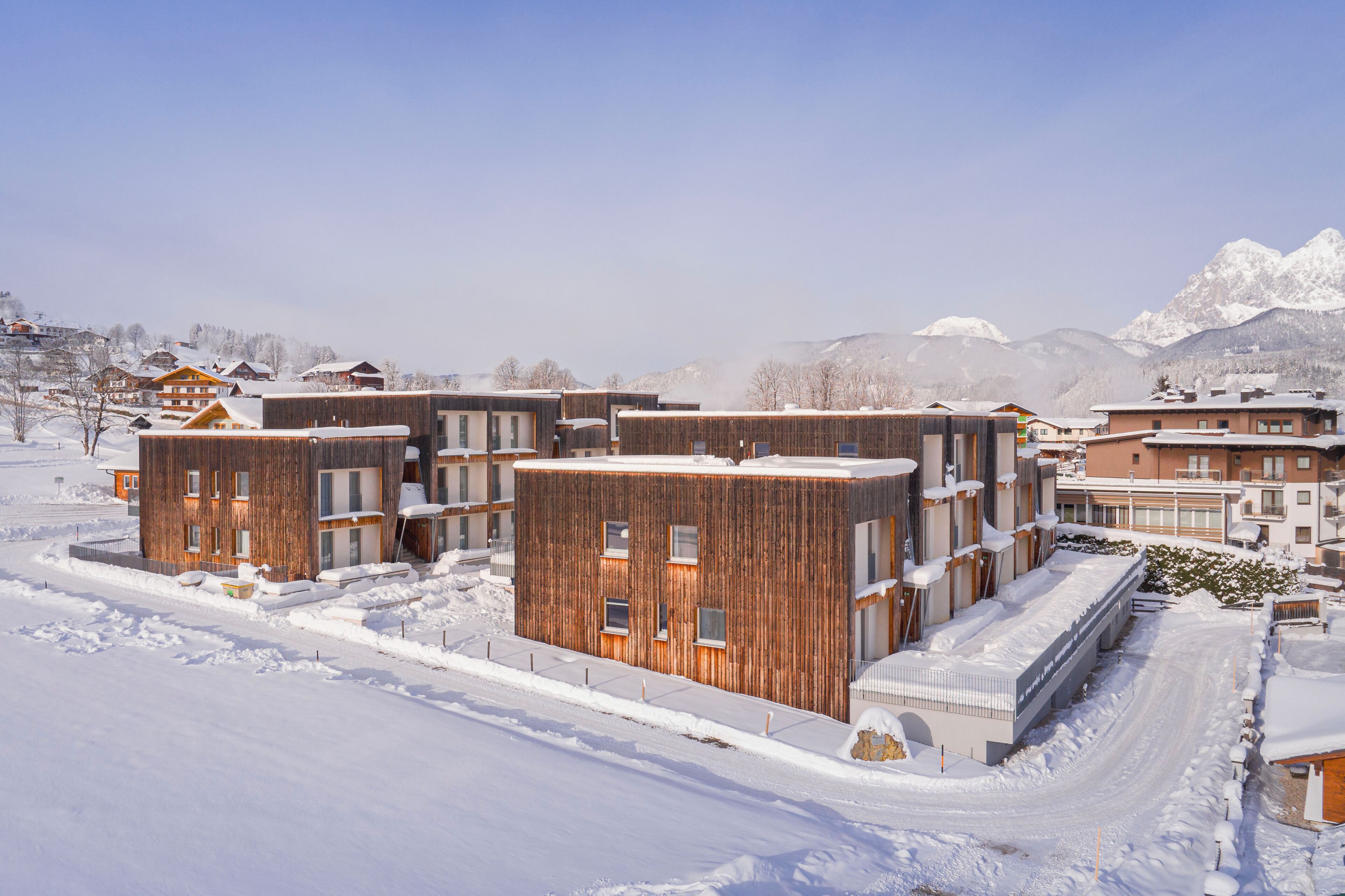 A modern building complex with multiple wooden facades, surrounded by snow-covered terrain and mountains in the background.