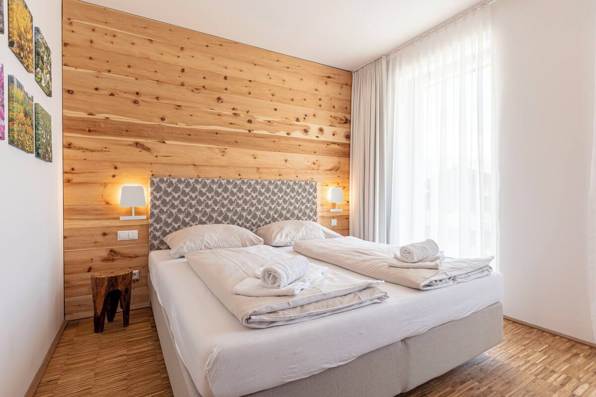 Een netjes opgemaakt bed met witte lakens en handdoeken, een houten hoofdbord met een patroon en een klein houten krukje ernaast.