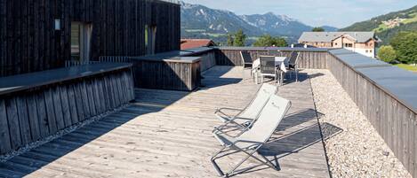 Una terraza de madera con vistas a montañas, un edificio de madera y una mesa con sillas.