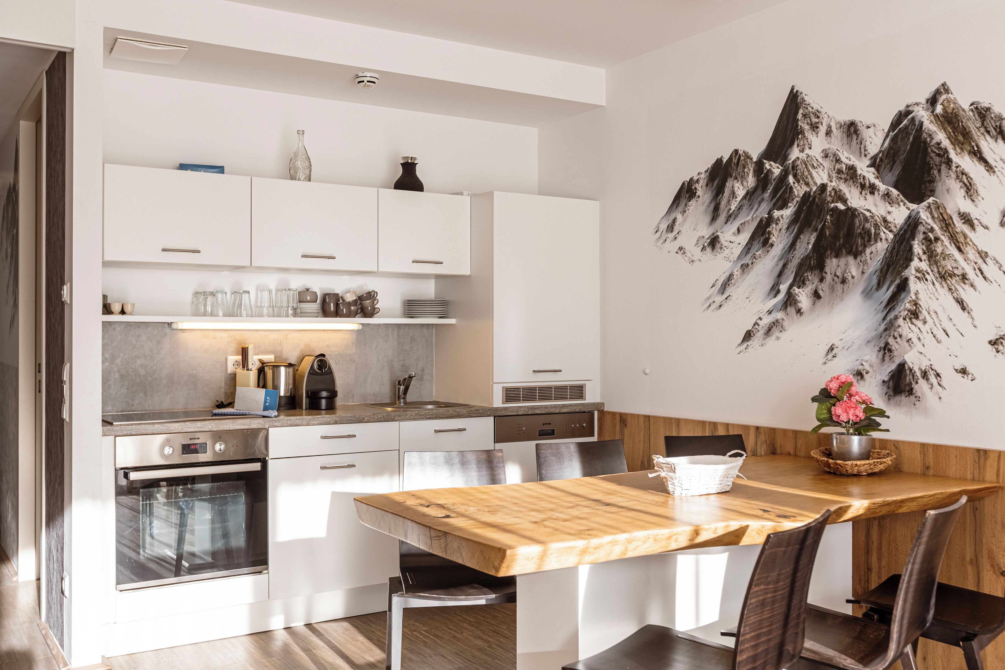 Une cuisine moderne avec des meubles blancs, une table à manger en bois et une fresque représentant une montagne sur le mur.