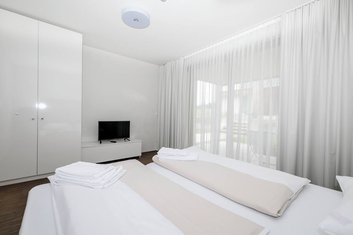 Een moderne hotelkamer met een groot bed, een televisie op een nachtkastje en uitzicht door dunne gordijnen.