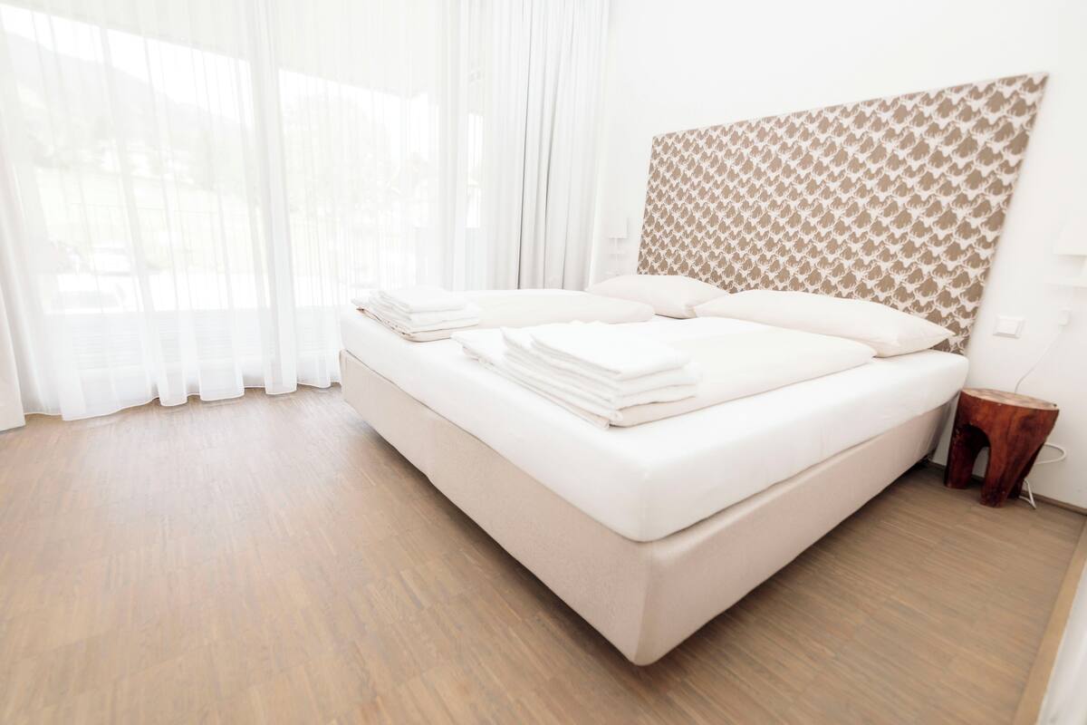 Een net opgemaakt bed met witte lakens in een kamer met houten vloeren en een kamerhoog hoofdbord.