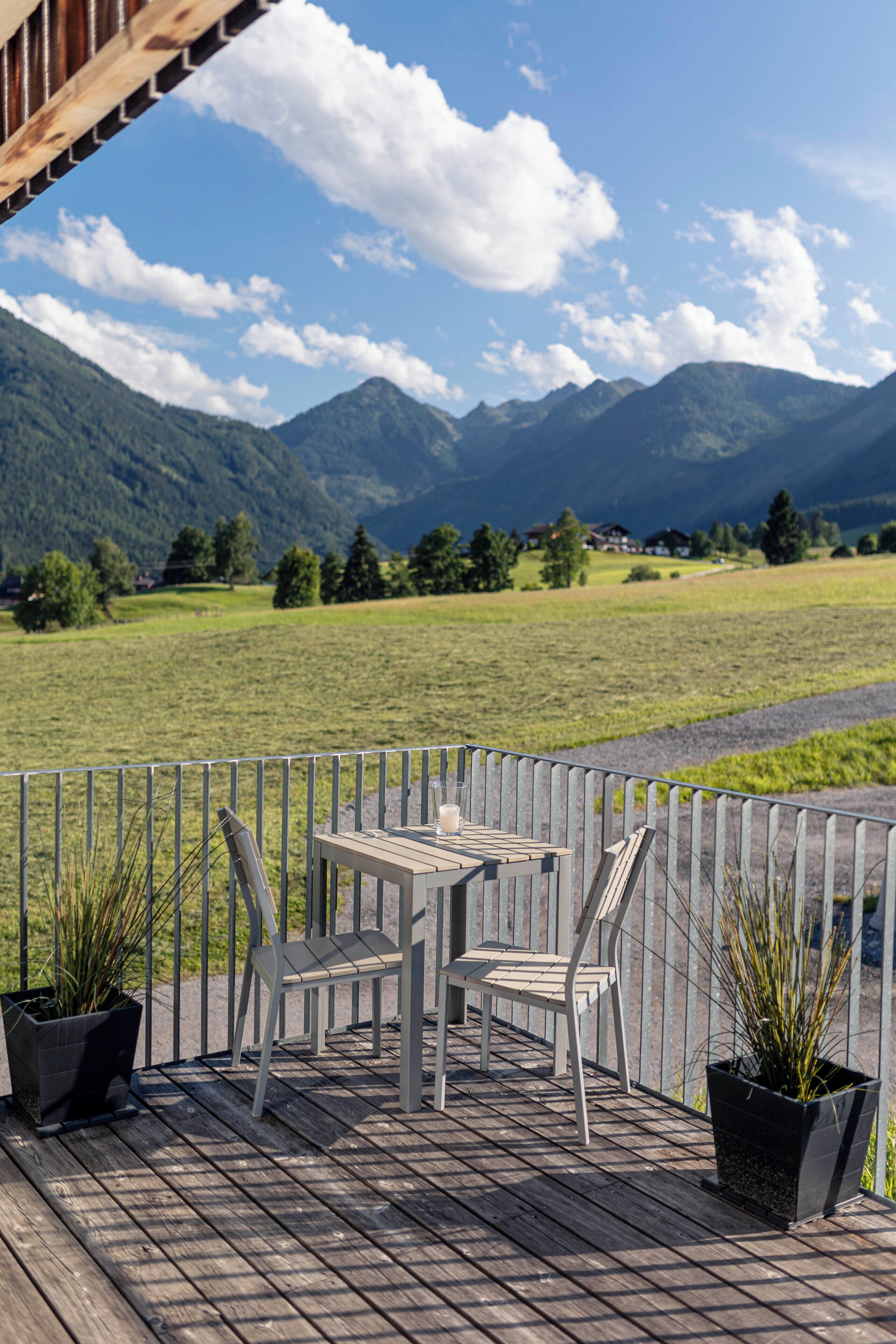 A wooden deck with a table and chairs, surrounded by a metal fence, overlooking a green field and mountains.