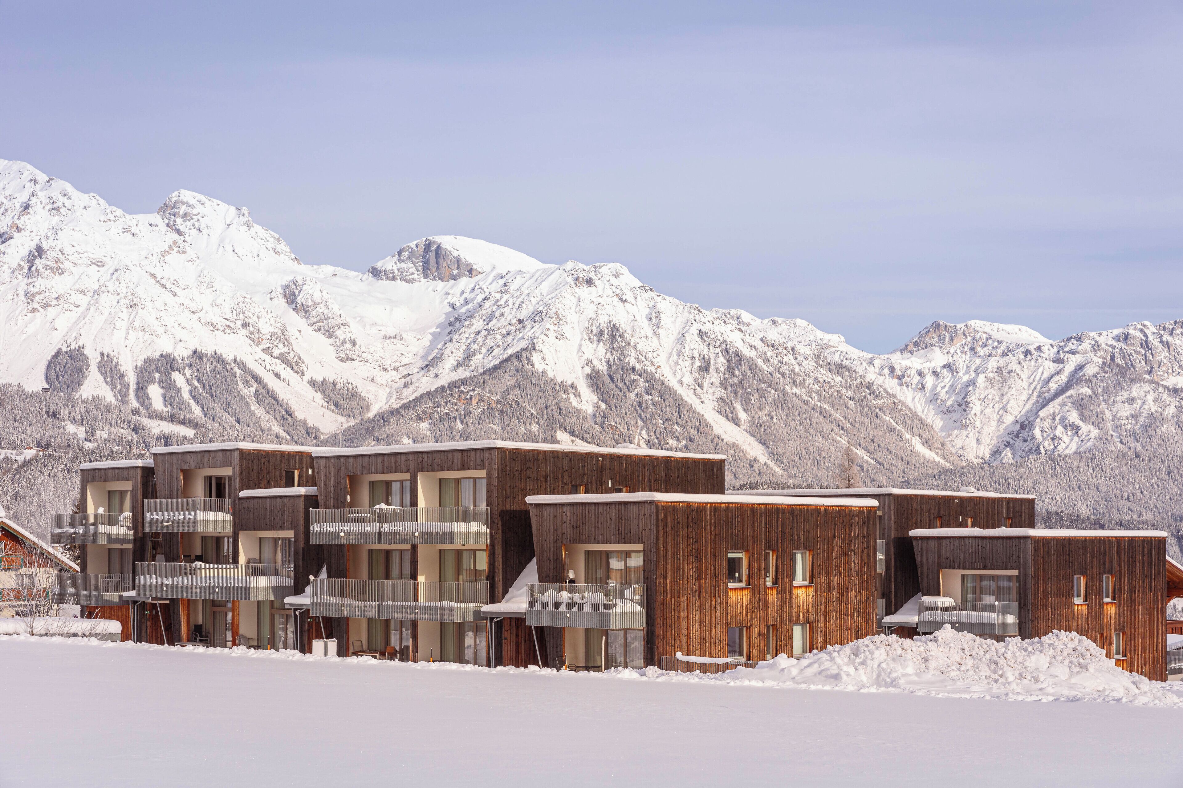 A modern building with multiple balconies and large windows, set against a snowy mountain backdrop.