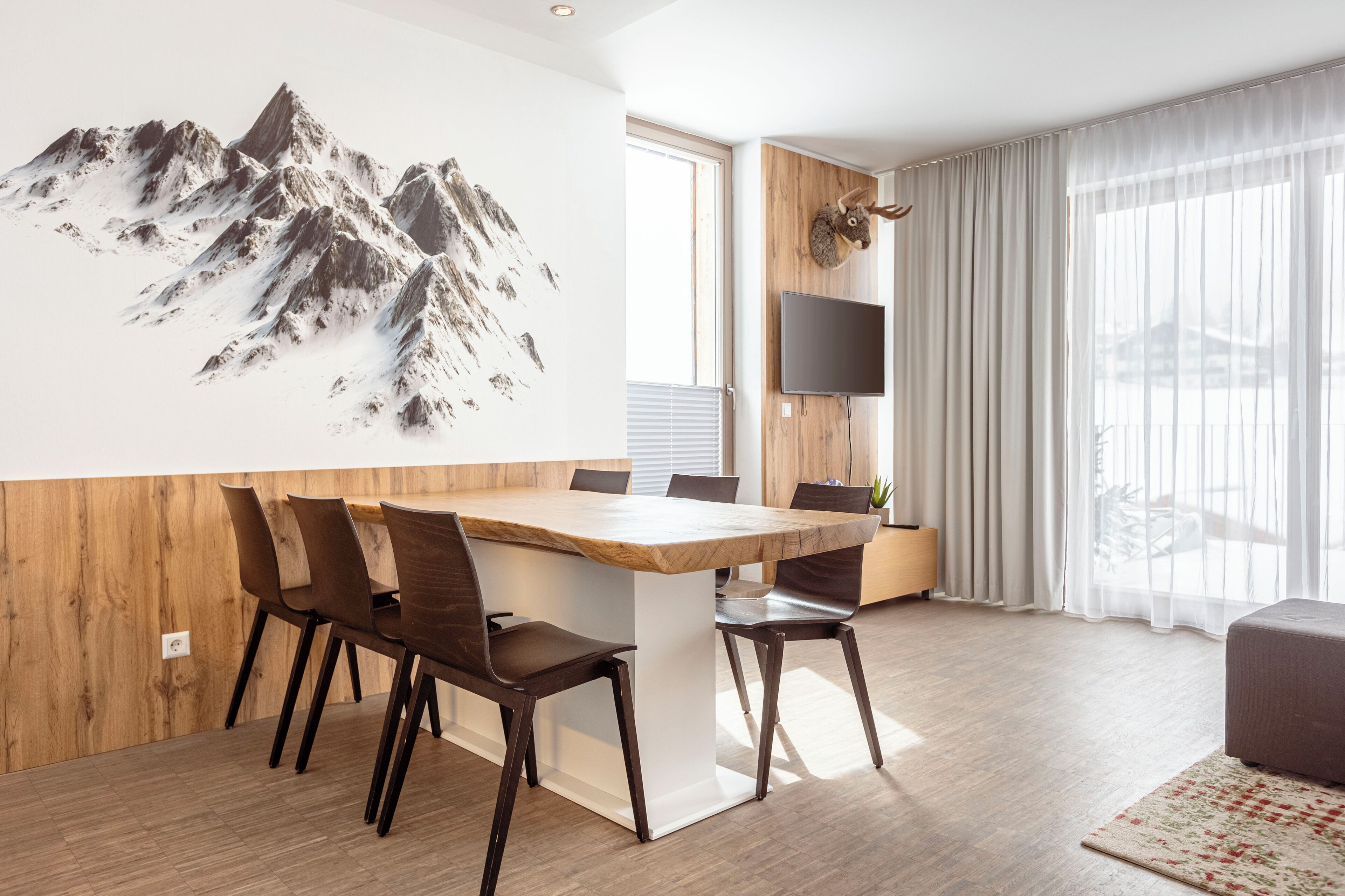 Un comedor moderno con una mesa y sillas de madera, un televisor montado y una obra de arte con motivos de montaña en la pared.