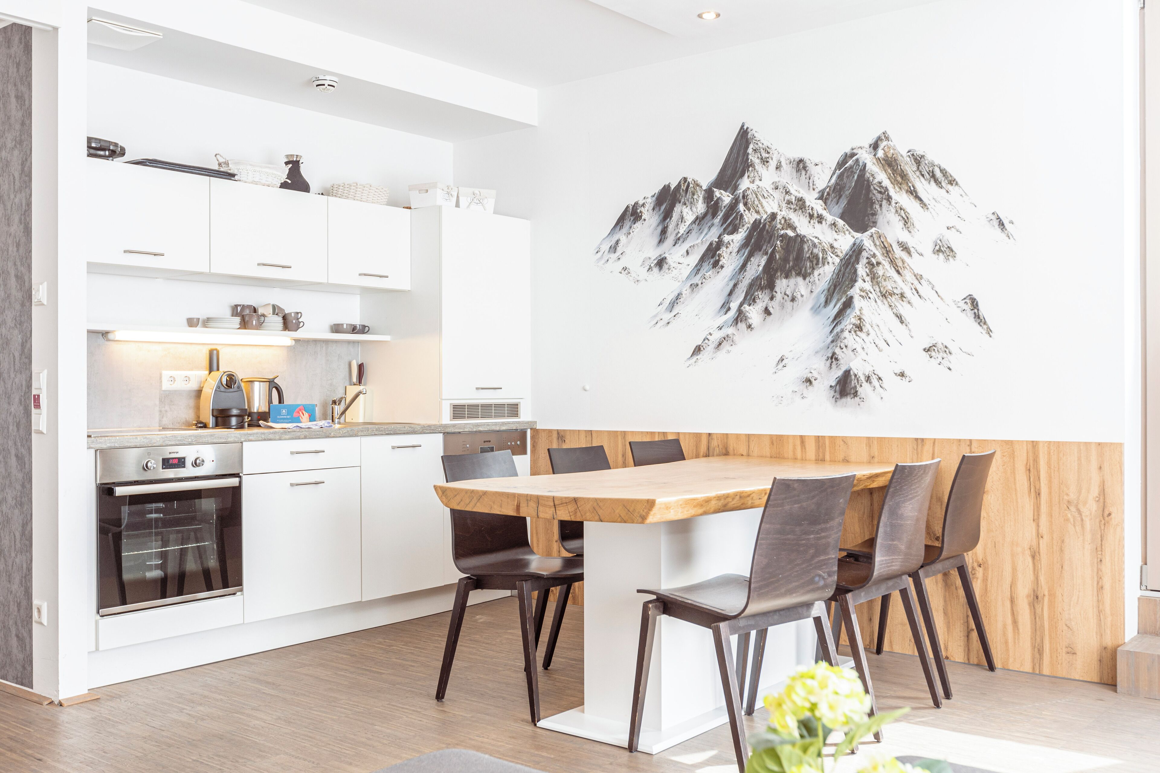 Une cuisine moderne avec des meubles blancs, une table à manger en bois et une fresque représentant une montagne sur le mur.