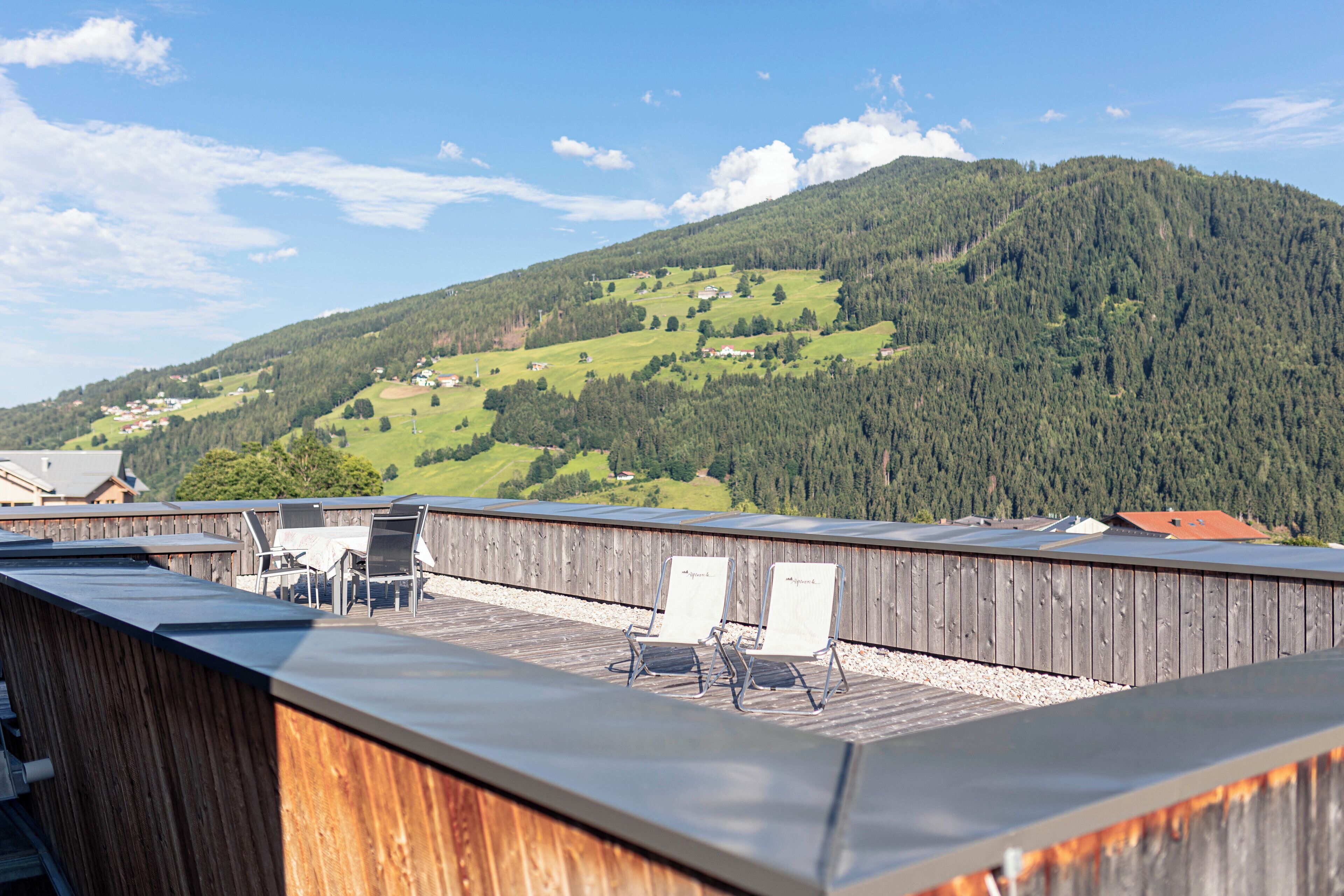 Una terraza de madera con dos sillas de lounge y una mesa, con vistas a un valle verde y colinas boscosas.