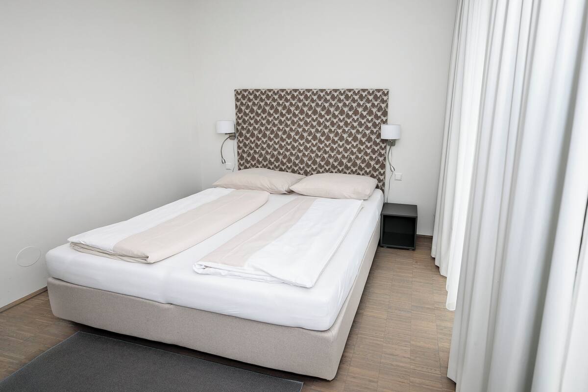 Een net opgemaakt bed met een gestreept hoofdbord, twee bedlampjes en uitzicht op een houten vloer en witte gordijnen.