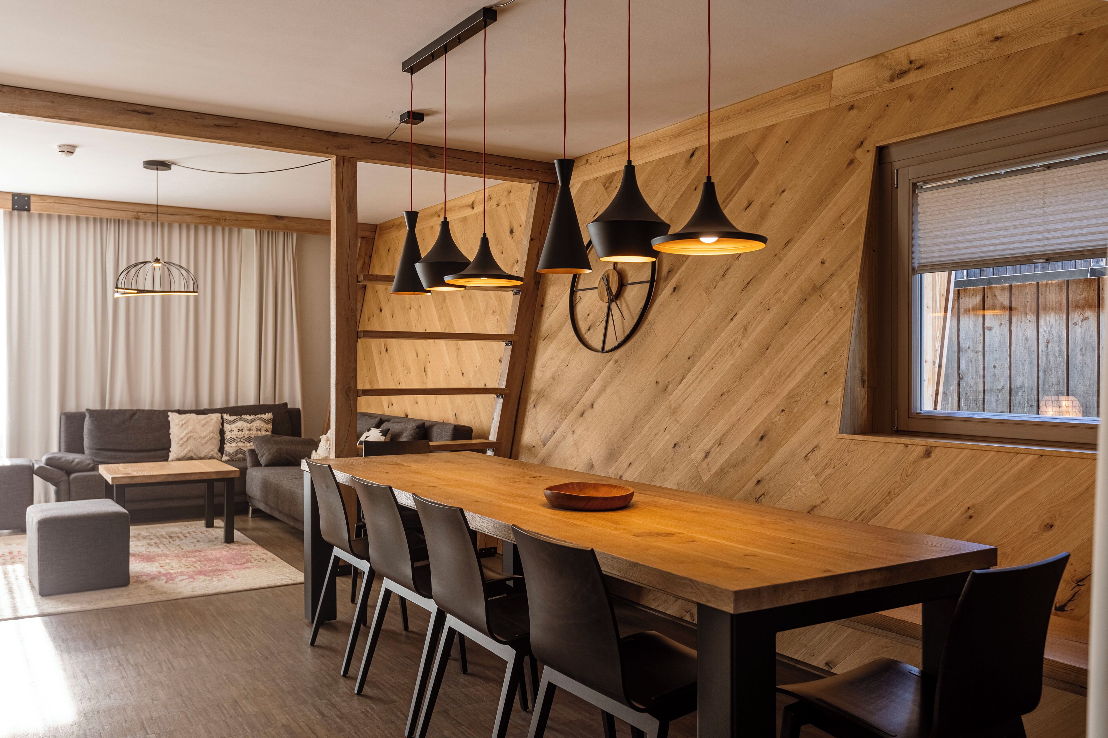 Un comedor moderno con una mesa y sillas de madera, luces colgantes y un amplio ventanal con persianas.