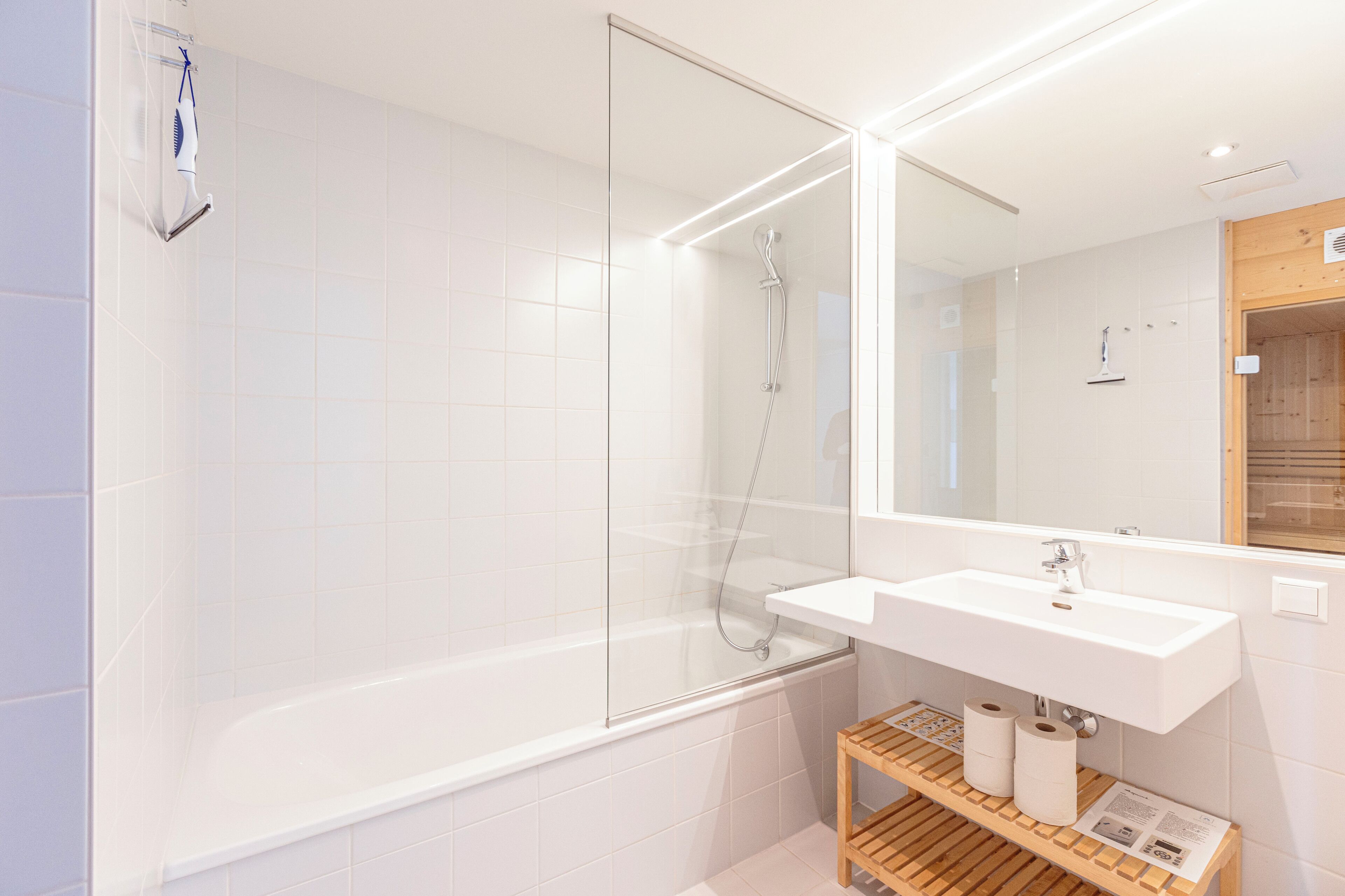 Une salle de bain moderne avec une douche à parois de verre, un lavabo blanc et une étagère en bois où sont rangés les articles de toilette.