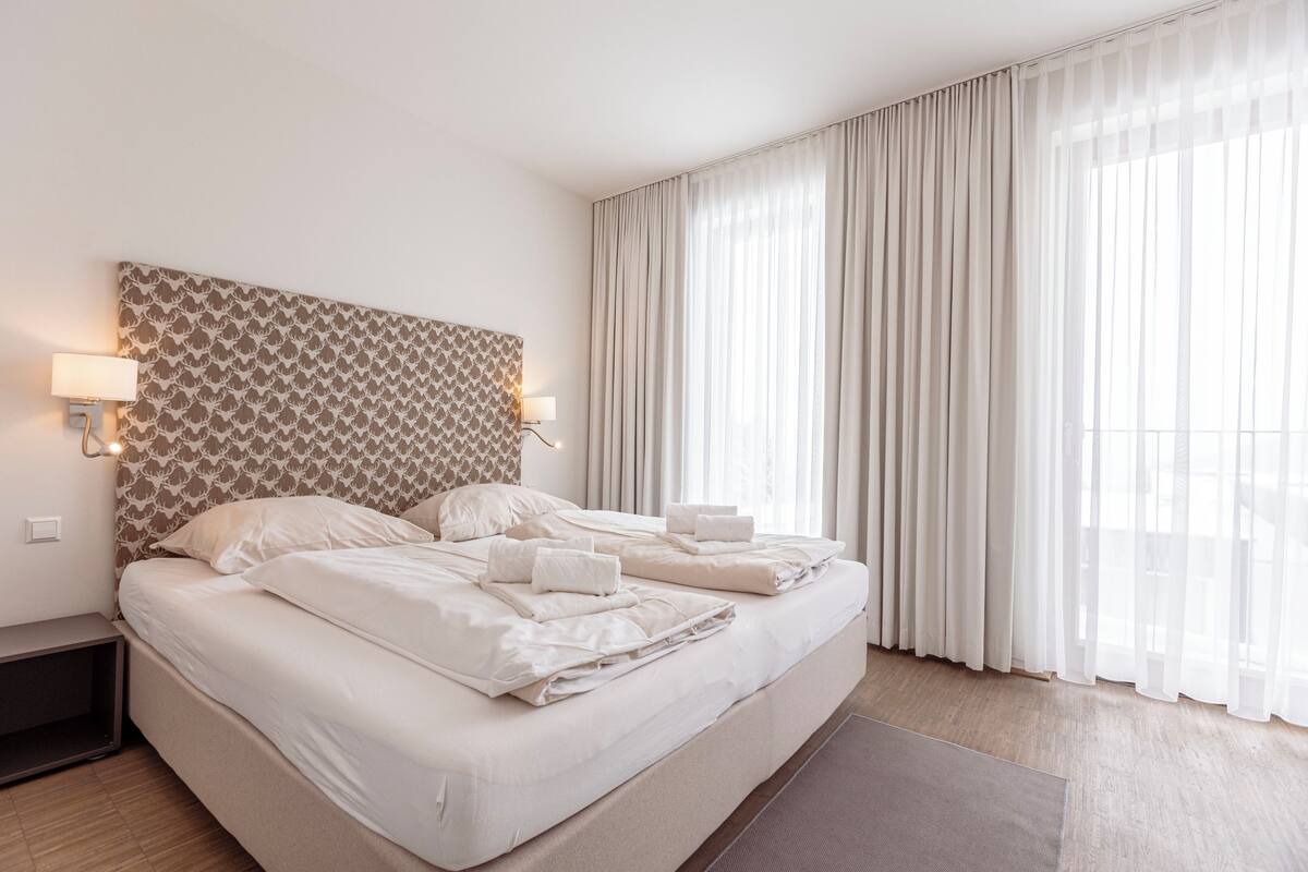 Een net opgemaakt bed met witte lakens en handdoeken, een hoofdbord met een patroon en een bedlampje.