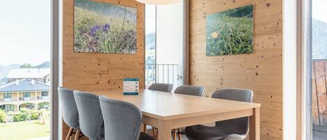A modern dining area with a wooden table and chairs, large windows with a view of buildings and greenery, and two framed botanical paintings on the walls.