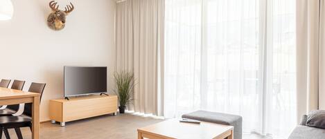 A modern living room with a wooden coffee table, a grey sofa, a TV on a wooden stand, a dining table with chairs, and a mounted deer head on the wall.