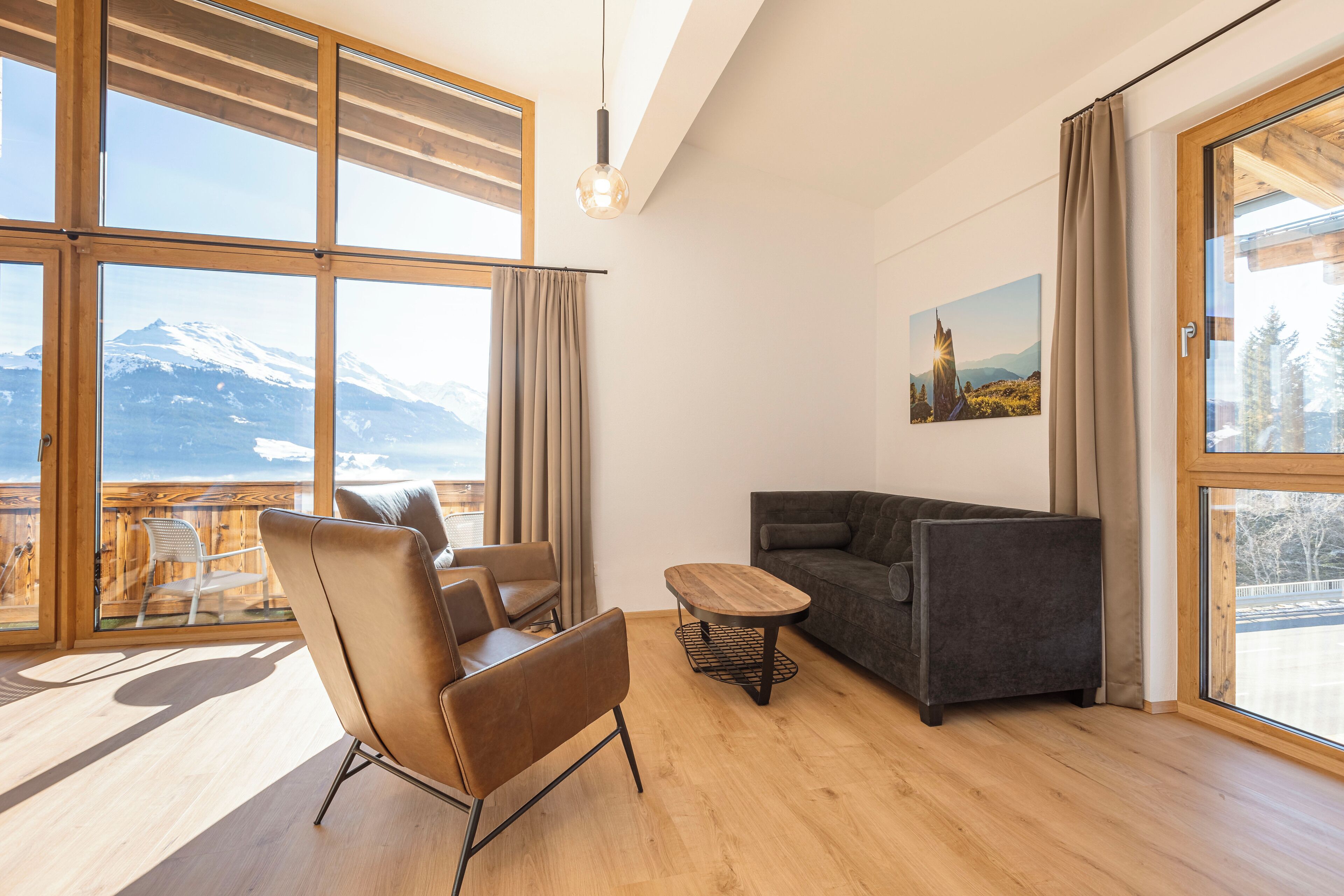 A modern living room with a wooden floor, a sofa, a chair, and a table. Large windows offer a view of mountains.