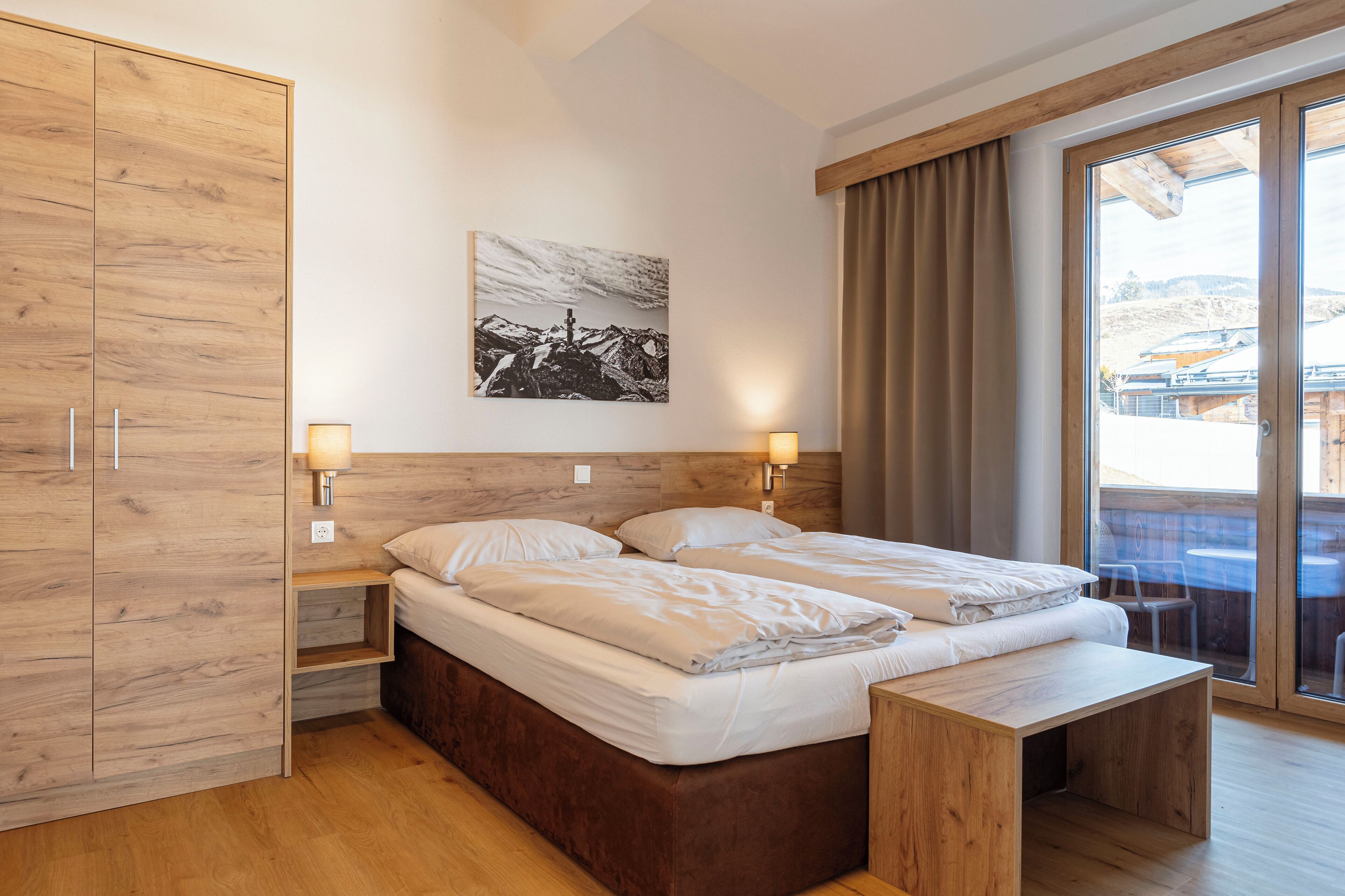 Ein modernes Hotelzimmer mit einem Holzbett, einem Nachttisch, einem Holzschrank und einem großen Fenster mit Blick nach außen.