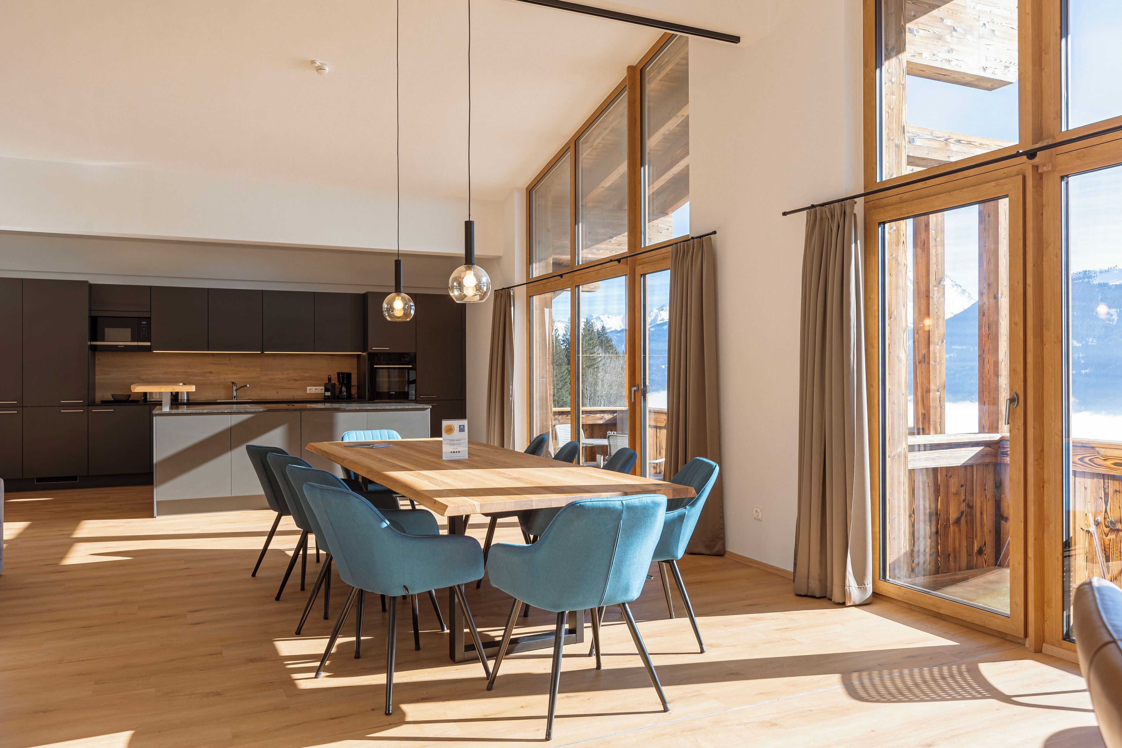 Une cuisine moderne avec des meubles de couleur sombre et un îlot central en bois, un coin repas avec une table en bois et des chaises bleues, et de grandes fenêtres offrant une vue sur les montagnes.