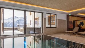 Ein moderner Innenpoolbereich mit großen Fenstern, Blick auf Berge und einer Lounge mit Sesseln.