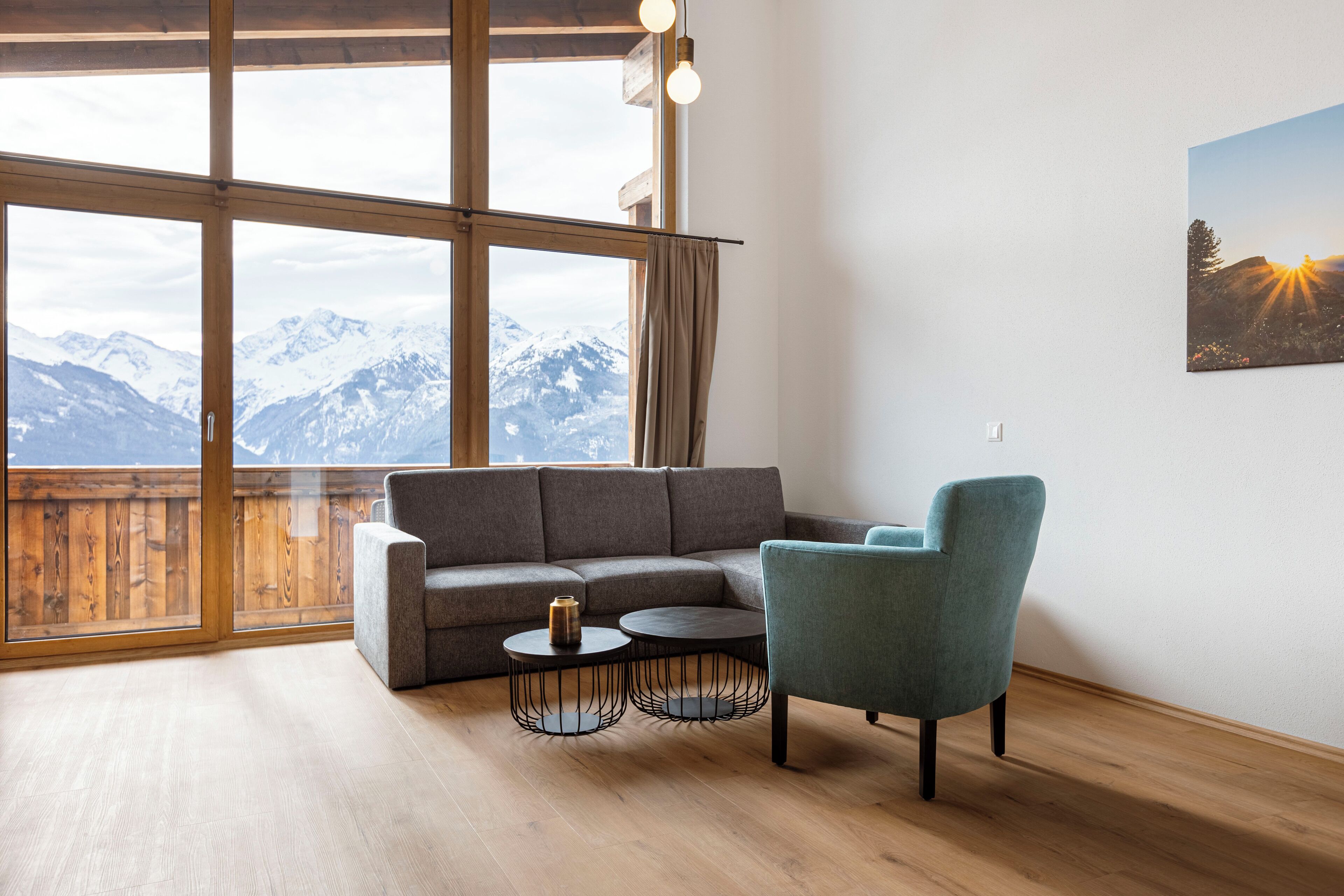 Una sala moderna con un sofá seccional gris, un sillón verde azulado y dos mesas de centro redondas. Un ventanal ofrece vistas de montañas nevadas.