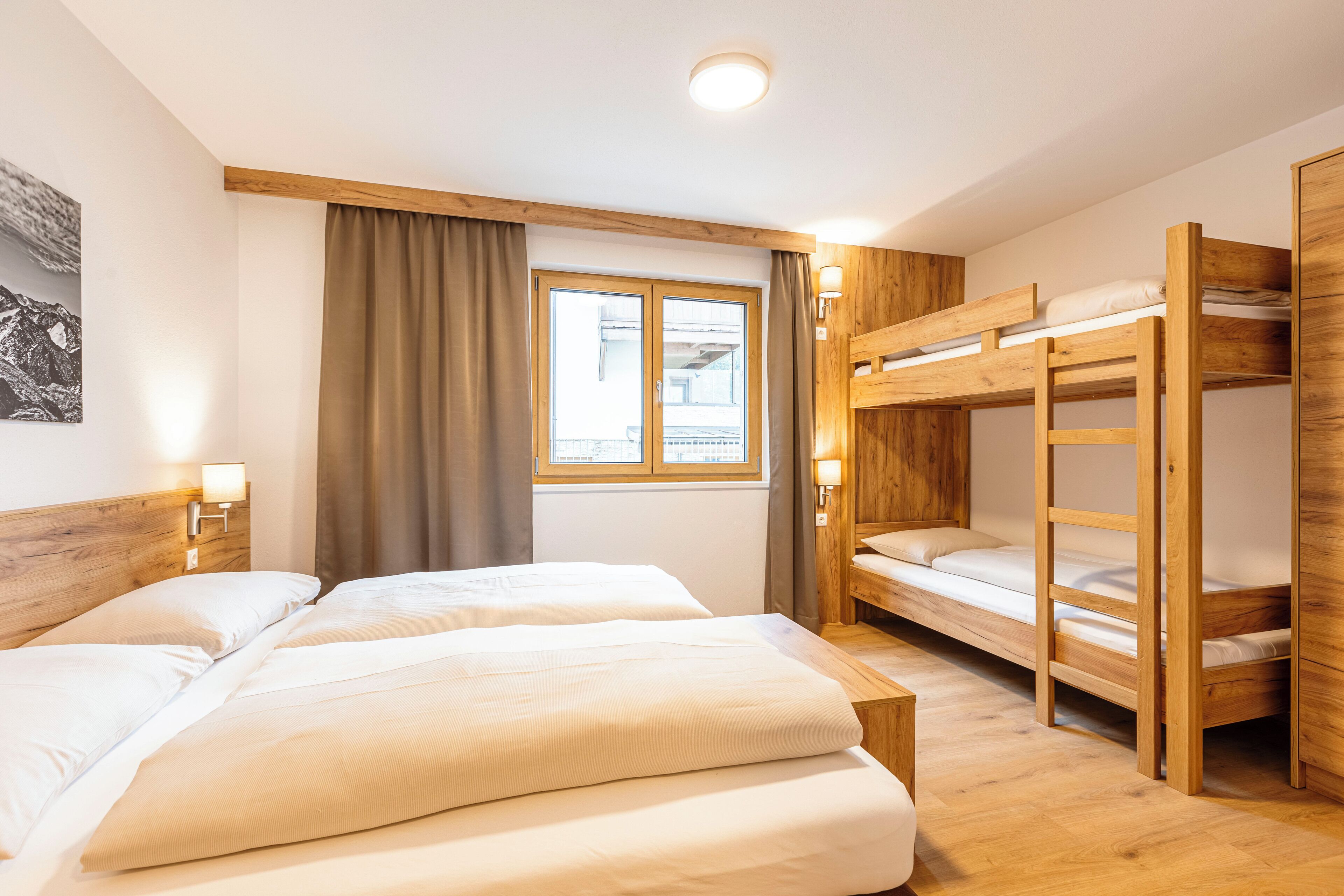 Een kamer met twee bedden, een stapelbed, een houten hoofdeinde, een raam met gordijnen en wandlampen.
