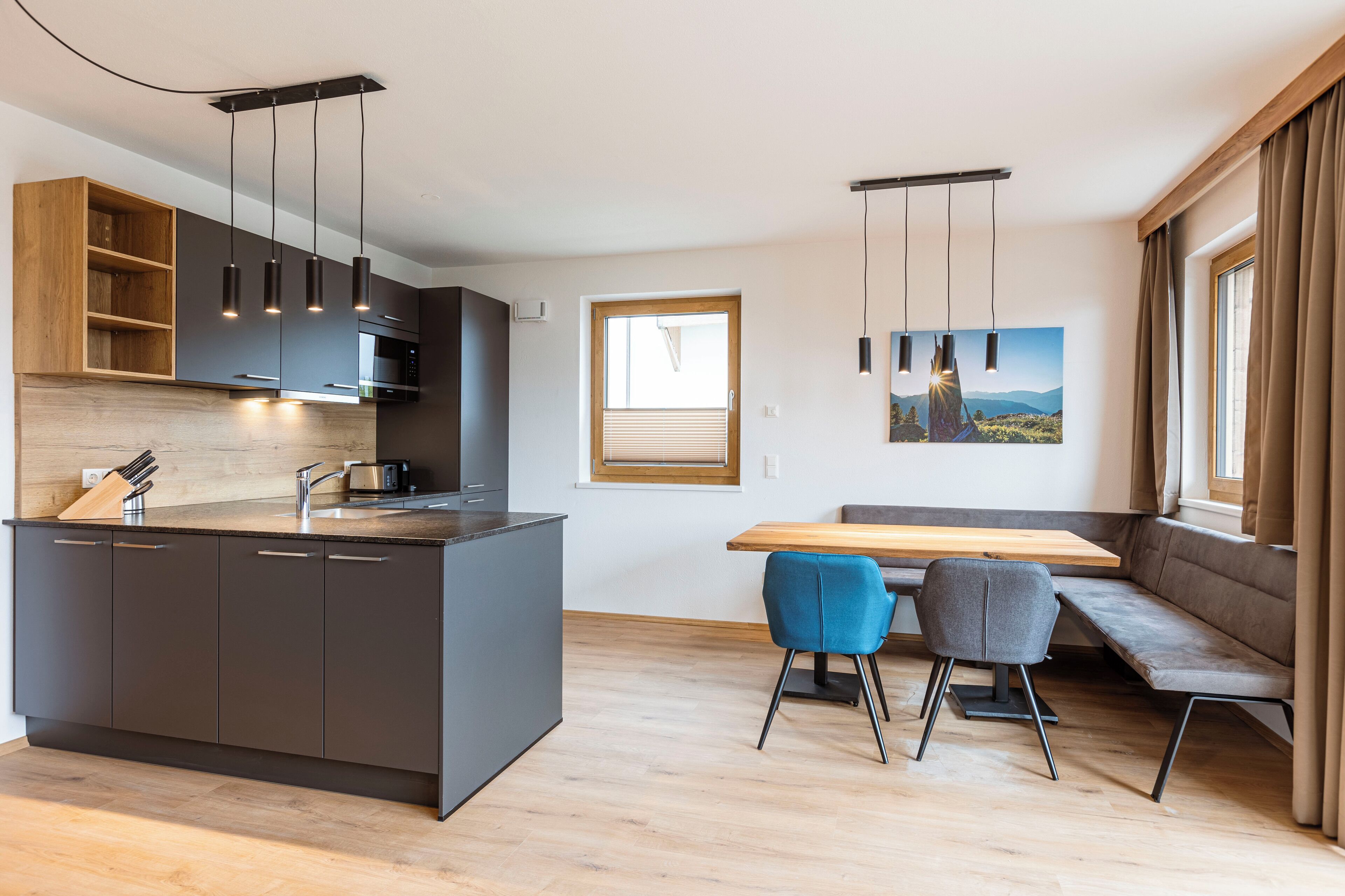 A modern kitchen with dark cabinetry, a wooden island, and pendant lighting. There is a dining area with a bench and two chairs.