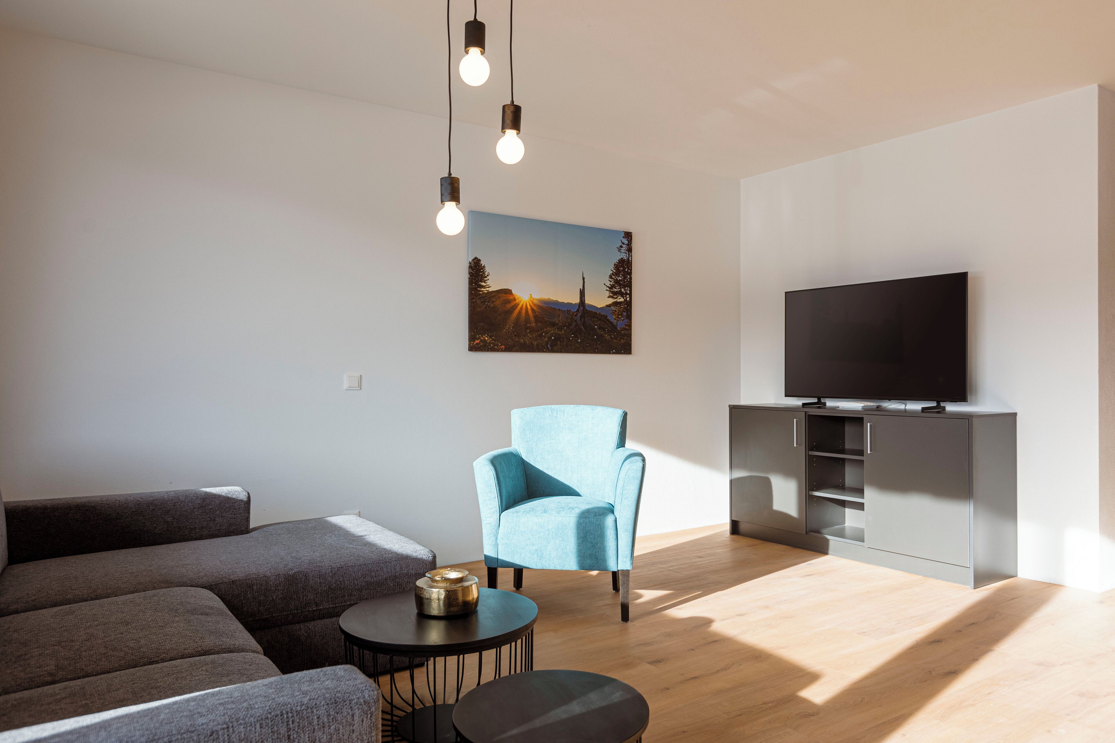 Ein modernes Wohnzimmer mit einem grauen Sofa, einem blauen Sessel, einem runden Couchtisch und einem Flachbildfernseher auf einem grauen Schrank.