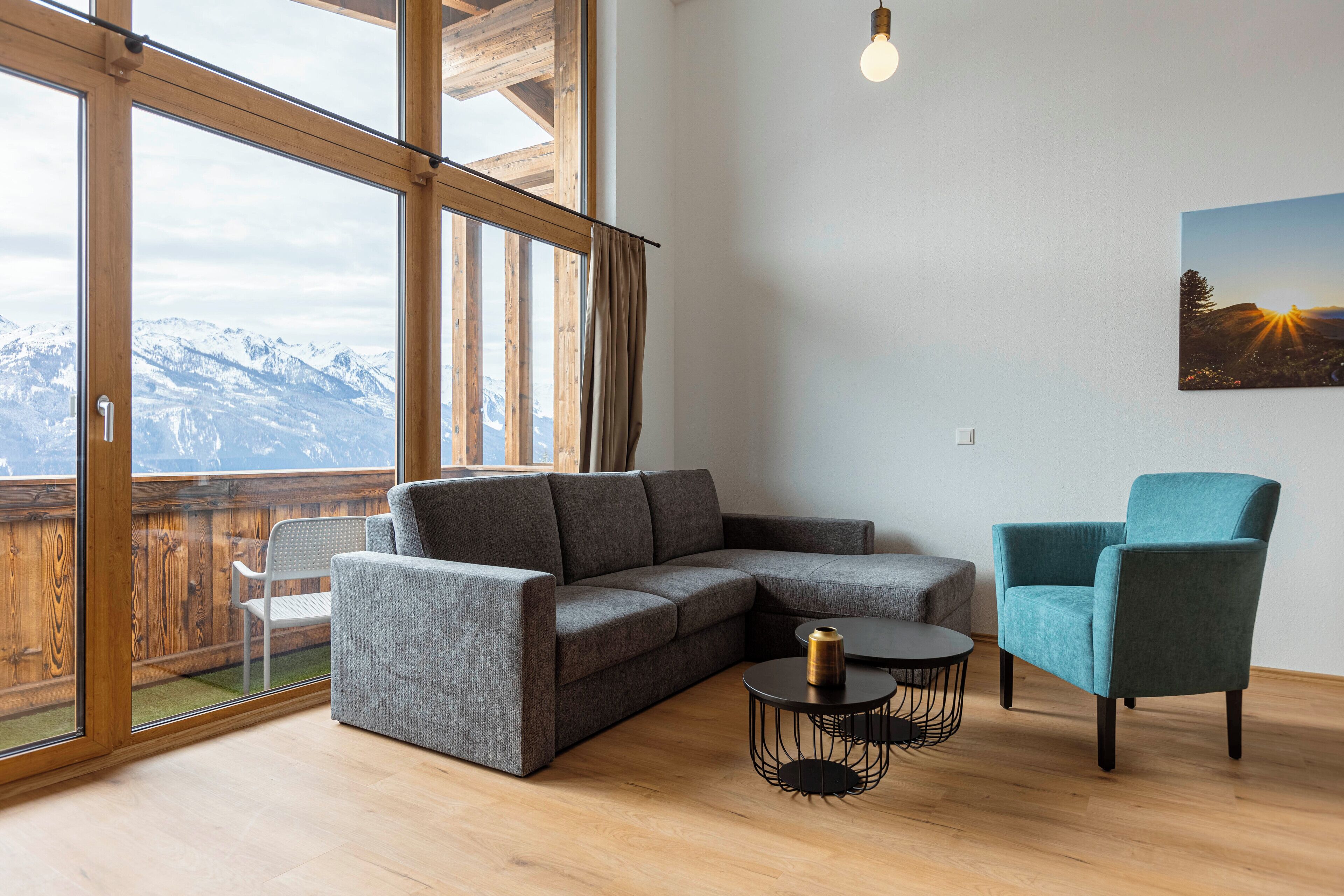 Uma sala de estar moderna com um sofá modular cinza, uma poltrona verde-água e uma pequena mesa redonda com luminária. Uma grande janela oferece vista para uma paisagem montanhosa.