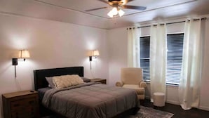 A bedroom with a bed, nightstands, a chair, a ceiling fan, and a window with curtains. - Amistad House (Del Rio)