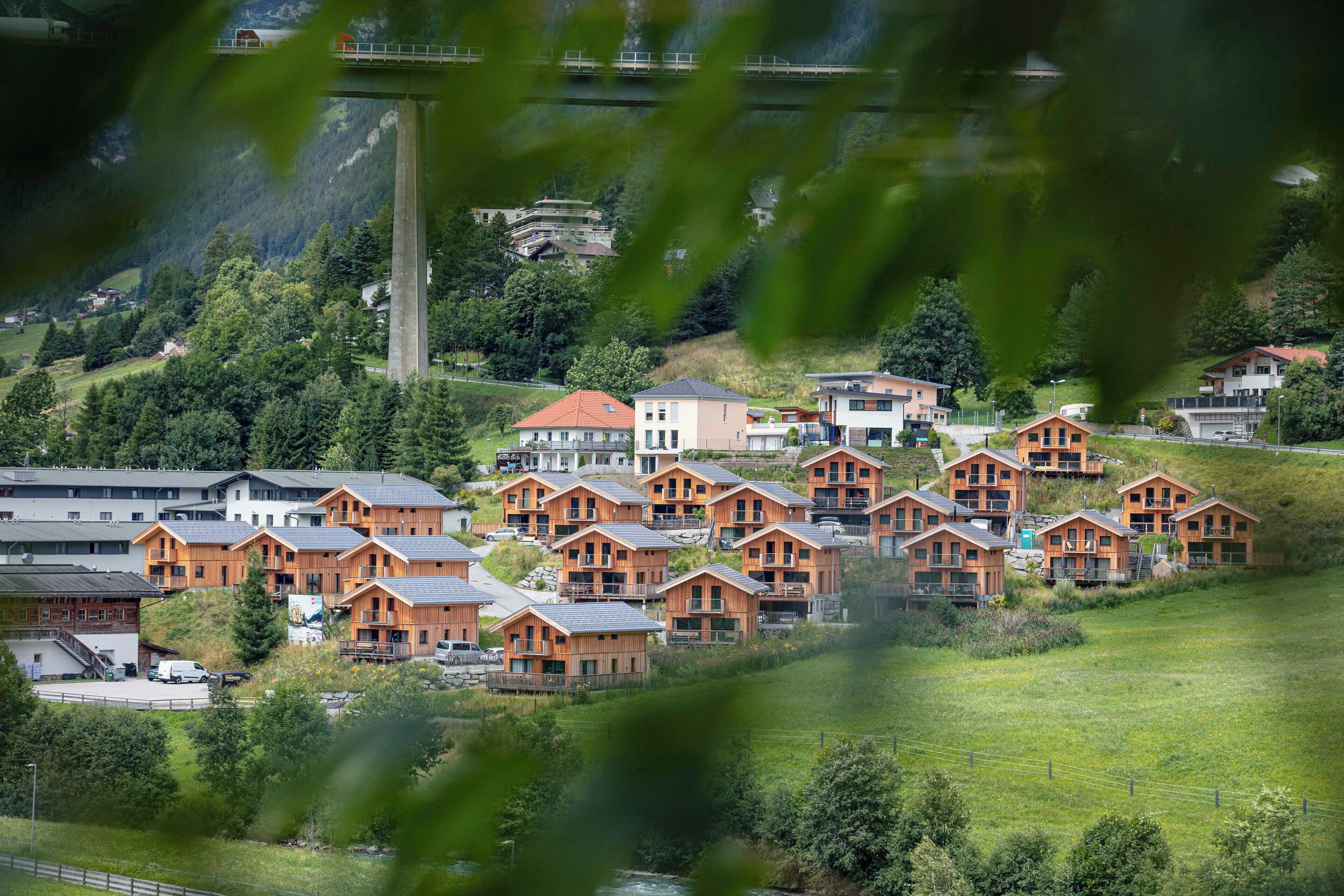 A cluster of wooden cabins with a mountain backdrop, a bridge, and a river in the foreground.