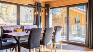 A modern interior with a wooden ceiling, large windows, a dining area with chairs and a table, and a sofa with cushions.