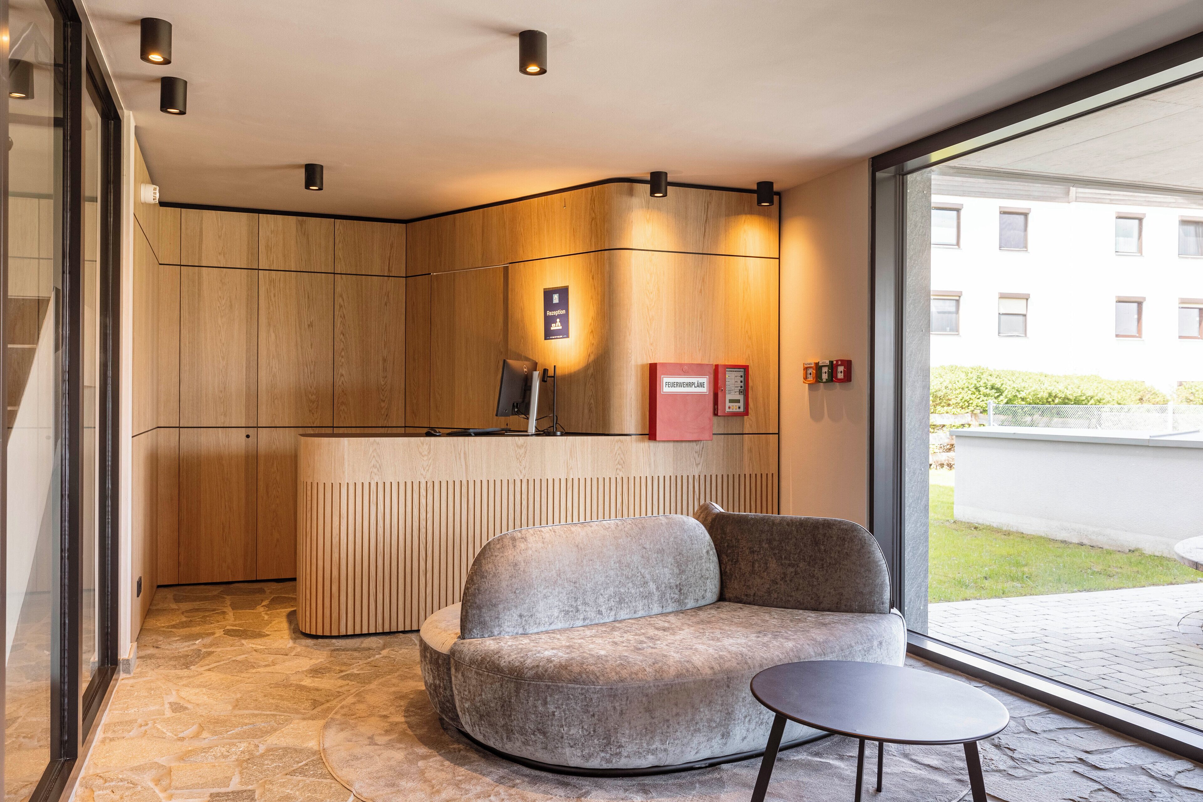 A reception desk with a wooden finish, a reception area with a sofa and a small table, and a view of a building outside.