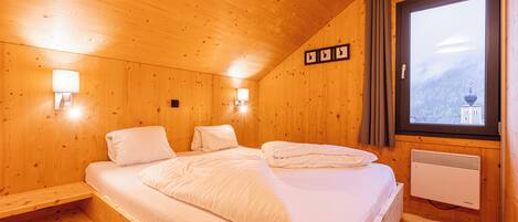 A wooden cabin bedroom with a bed, two wall-mounted lamps, a window with curtains, and a radiator.