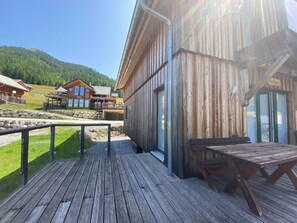 Une terrasse en bois avec une table et des chaises, un mur en pierre et une vue sur une montagne et des maisons au loin.