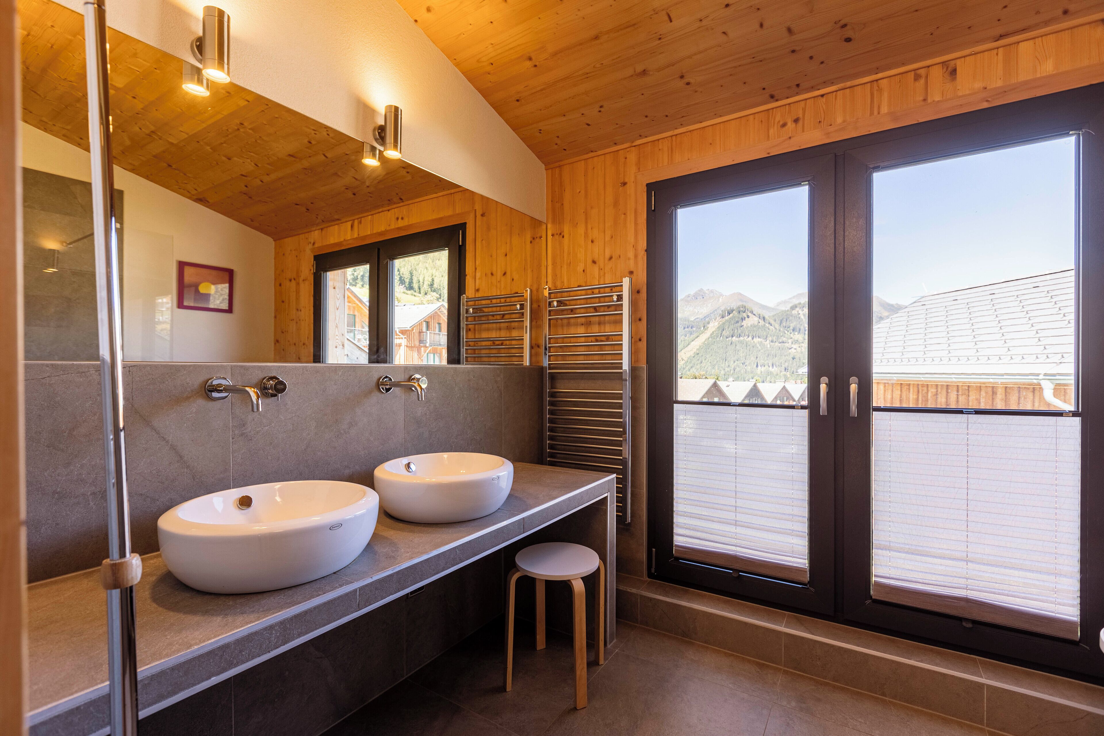 A modern bathroom with two sinks, a large mirror, and a view of mountains through the windows.