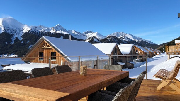 Ein Holzespeisetisch mit Korbstühlen in einer verschneiten Winterlandschaft, umgeben von Blockhütten mit schneebedeckten Dächern und Bergen im Hintergrund.