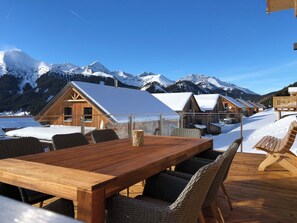 Une table à manger en bois, entourée de chaises en osier, dans un décor extérieur enneigé, avec des chalets aux toits couverts de neige et des montagnes en arrière-plan.