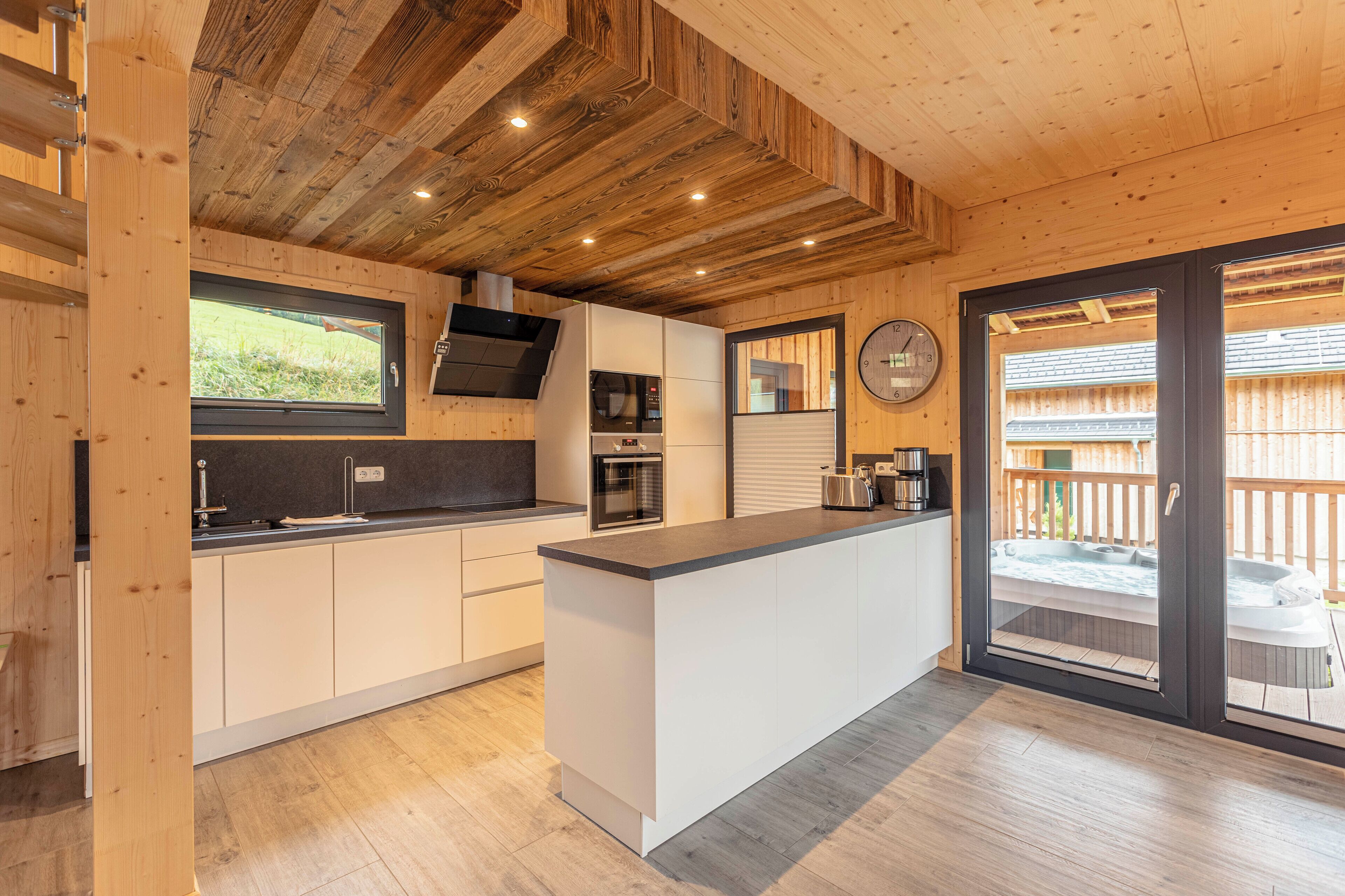 A modern kitchen with wooden ceiling beams, a large island, built-in appliances, and a view of a hot tub outside.