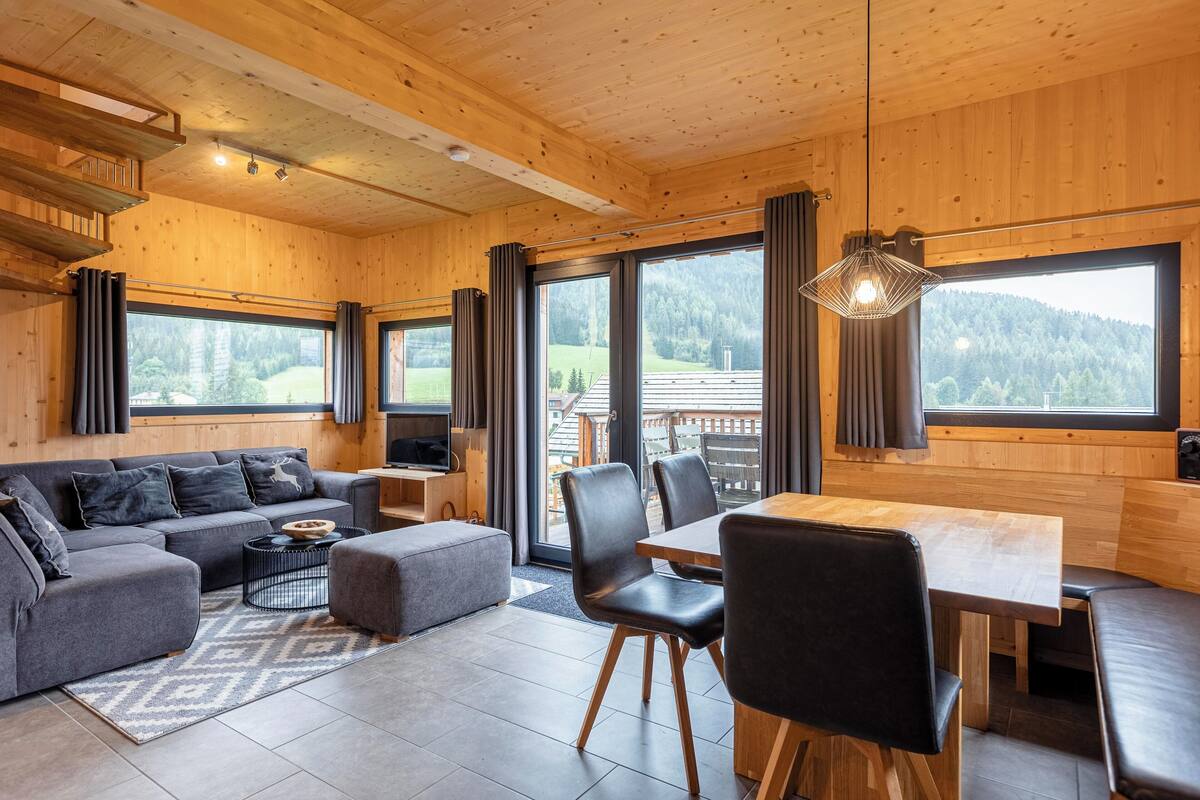 Un interior moderno con techo de madera, un sofá gris, una mesa de comedor de madera y grandes ventanales que dan a una zona boscosa.
