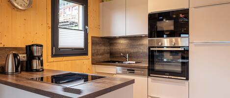 A modern kitchen with a built-in stove, oven, and dishwasher. There is a window with blinds, a clock on the wall, and a coffee maker on the counter.