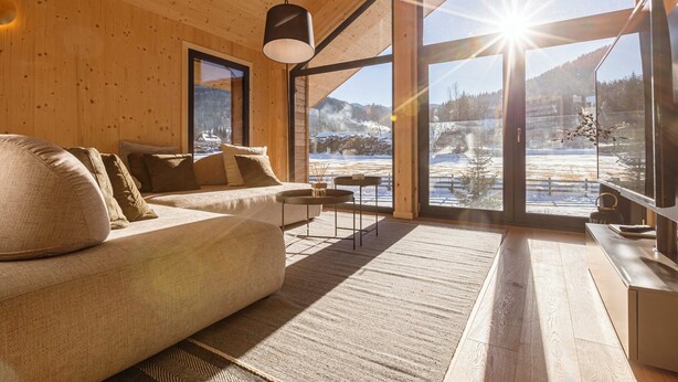 A cozy cabin interior with a wooden interior, a sofa, a coffee table, and a large window offering a snowy mountain view.